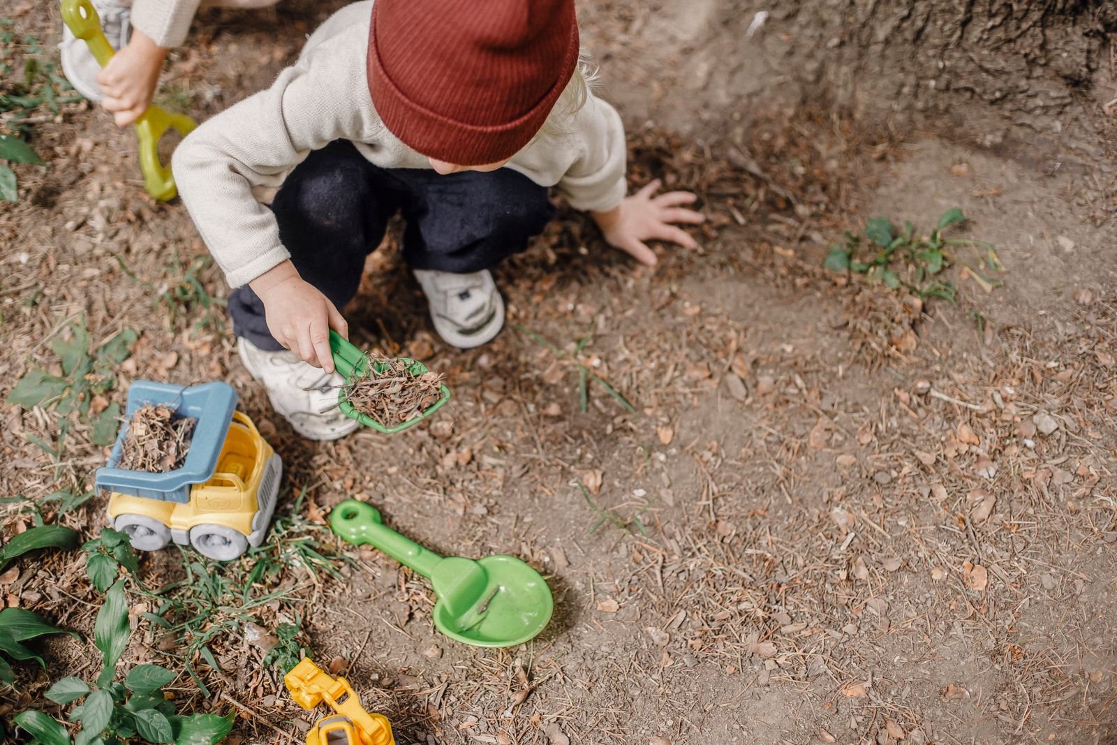 A young child crouching in a garden, digging with a toy spade beside a toy truck
